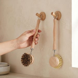 May include: Two wooden dish brushes with natural bristles hanging on a wall. The brushes have long, slender handles and round brush heads. One brush is being held by a hand. The wall hooks are made of wood and have a simple, minimalist design.