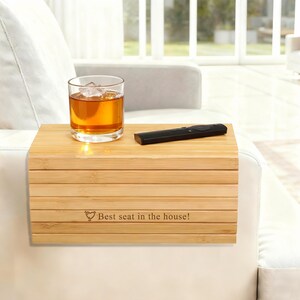 May include: A bamboo sofa arm tray with a glass of amber liquid and ice, and a black remote control. The tray has the text "Best seat in the house!" engraved on it. The tray is designed to rest on a sofa arm.