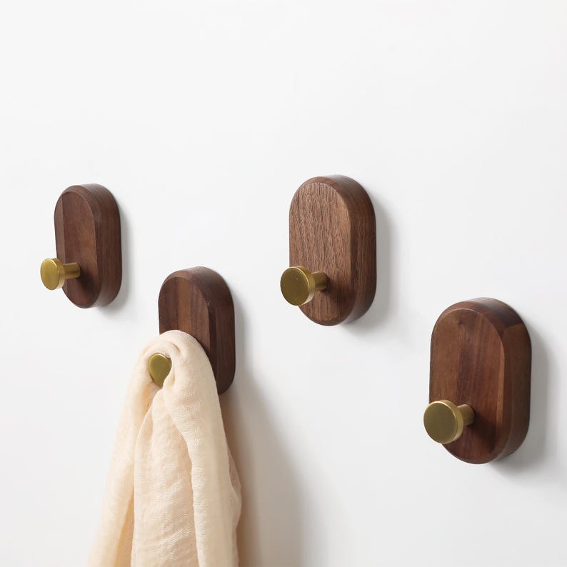 May include: Four wooden wall hooks with brass knobs. The hooks are oval-shaped and made of dark brown wood. One hook holds a cream-colored scarf. The hooks are mounted on a white wall.