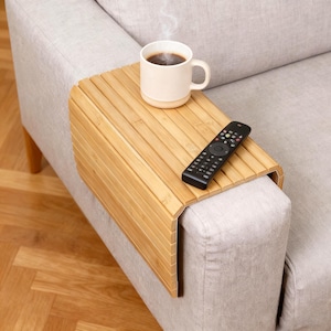 May include: A bamboo sofa arm tray with a cup of coffee and a remote control. The light brown tray is made of parallel bamboo slats and rests on the arm of a light gray sofa. A white mug with coffee sits on the tray, and a black remote control is next to it.