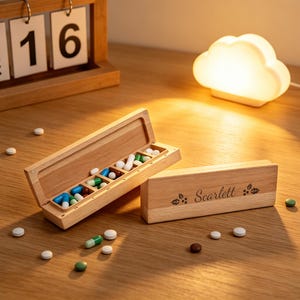 Op de afbeelding: Een houten pillenorganizer met de naam "Scarlett" gegraveerd op het deksel. De open organizer toont verschillende pillen en capsules in verschillende kleuren. Een houten kalender en een wolkvormige lamp staan op de achtergrond.