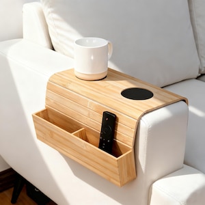 May include: A bamboo sofa arm tray with a built-in cup holder and storage compartments. A white mug sits on the tray, and a black remote is in the storage. The tray is designed to fit over a sofa arm.