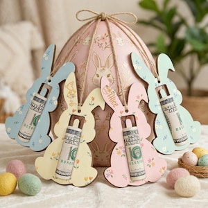 May include: Wooden Easter bunny gift holders in pastel colors, each holding a rolled-up bill. The bunnies are light blue, yellow, and pink, decorated with floral patterns. A large pink egg-shaped box with bunny illustrations is in the background.
