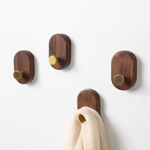 May include: Four wooden wall hooks with brass knobs. The hooks are oval-shaped and made of dark brown wood. One hook holds a light beige scarf. The hooks are mounted on a white wall, offering a minimalist and functional design.