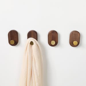 May include: Four wooden wall hooks with brass knobs are mounted on a white wall. One hook holds a cream-coloured scarf. The hooks are oval-shaped and made of dark wood. The brass knobs add a touch of elegance to the design.