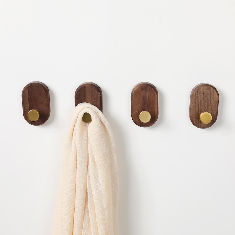 May include: Four wooden wall hooks with brass knobs are mounted on a white wall. One hook holds a cream-coloured scarf. The hooks are oval-shaped and made of dark wood. The brass knobs add a touch of elegance to the design.