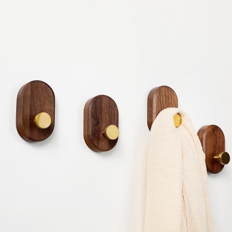May include: Four wooden wall hooks with brass knobs are mounted on a white wall. The hooks are oval-shaped and made of dark brown wood. One hook holds a cream-coloured scarf. The brass knobs add a touch of elegance to the minimalist design.