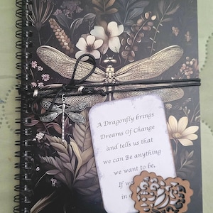 May include: A spiral-bound notebook with a dark floral cover featuring a large dragonfly illustration. A black cord wraps around the cover, holding a silver dragonfly charm and a white card with the text: A Dragonfly brings Dreams Of Change and tells us that we can Be anything we want to be, If we in o. A brown heart-shaped cutout is attached.