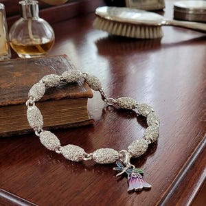 May include: A silver-toned bracelet composed of oval-shaped links covered in clear crystals. The bracelet features a dangling charm shaped like a flower with purple and blue enamel petals and a small dragonfly accent attached to the top of the bloom.