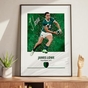 May include: Framed print featuring a rugby player in a green jersey, holding a ball. The print has a green and white background with the player's signature and name, "James Lowe," and team information. The artwork is displayed on a wooden surface.
