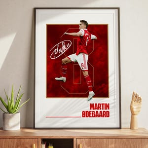 May include: Framed print featuring a soccer player in a red and white uniform, with the number 8. The player is positioned on a large number 8 with a red background. The print includes the player's signature and the name "MARTIN ØDEGAARD" in bold, red letters.