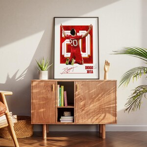 Pode incluir: Um poster emoldurado com um jogador de futebol de uniforme vermelho, ajoelhado com os bra&ccedil;os levantados. O poster tem um design vermelho e branco, o n&uacute;mero 20 e uma assinatura. O texto "DIOGO JOTA" tamb&eacute;m &eacute; vis&iacute;vel.
