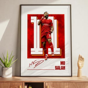 May include: Framed poster featuring a soccer player in a red uniform with the number 11. The player is celebrating a goal. The poster has a red and white background with the player's name, "MO SALAH", and signature.