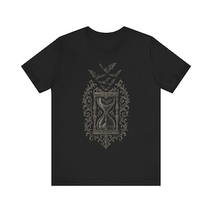 May include: Black t-shirt featuring a detailed graphic of an hourglass, bats, and ornate scrollwork. The design is in shades of gray and beige. The shirt is a short-sleeved crew neck.