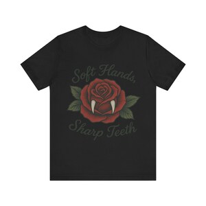 May include: Black t-shirt featuring a red rose with vampire fangs and green leaves. The text "Soft Hands, Sharp Teeth" is arched above and below the rose. The design has a vintage, tattoo-inspired aesthetic.