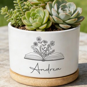 May include: A white cylindrical ceramic planter with a bamboo saucer base, containing several green succulents. The front of the planter features a dark gray graphic of an open book with flowers growing out of it and the name Andrea printed below.