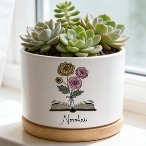Pode incluir: Um vaso cilíndrico de cerâmica branca com base de madeira clara, contendo suculentas verdes. A frente apresenta um gráfico de um livro aberto com um buquê de flores amarelas e rosas, com o nome Novalee escrito logo abaixo.