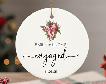 Puede incluir: Adorno navideño redondo de cerámica blanca con diseño floral y lazo. El adorno tiene las palabras "Emily + Lucas" y "engaged" con la fecha "11.08.25". Se adjunta un cordón dorado para colgarlo.