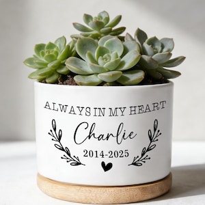 Puede incluir: Una maceta de cerámica blanca con una planta suculenta. La maceta tiene el texto "ALWAYS IN MY HEART" sobre el nombre "Charlie" y los años "2014-2025", con un símbolo de corazón. Se asienta sobre una base de madera.