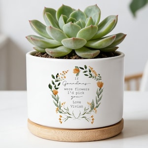 Könnte beinhalten: Weißer Keramik-Blumentopf mit einer Sukkulente. Der Topf steht auf einem Holzsockel und hat ein Blumengirlanden-Design mit dem Text "If Grandmas were flowers I'd pick you Love Vivian."