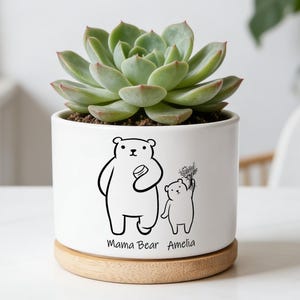 Könnte beinhalten: Ein weißer Keramik-Pflanztopf mit einer Sukkulente. Der Topf zeigt eine schwarz-weiße Illustration einer Mama-Bärin und eines Baby-Bären mit dem Text "Mama Bear Amelia". Er steht auf einem runden Holzsockel.