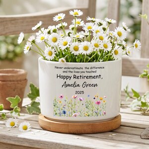 May include: A white ceramic planter with a bamboo base, filled with white daisy flowers. The planter features the text "Never underestimate the difference and the lives you touched Happy Retirement, Amelia Green 2025" above a colorful floral graphic.