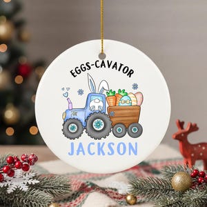 以下が含まれることがあります： 白いセラミックイースターオーナメント。青いトラクターのデザインで、ウサギの耳が付いたトラクターが、イースターエッグとニンジンを積んだカートを引っ張っています。「Eggs-cavator」と「Jackson」の文字がプリントされています。