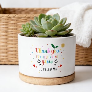 May include: A white ceramic plant pot with a succulent, inscribed with "Thank you for helping me grow, Love, Emma" in colourful text. The pot rests on a wooden base, with a woven basket and a white towel in the background.