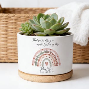 Puede incluir: Una maceta de cerámica blanca con base de madera, con una planta suculenta. La maceta tiene un diseño de arcoíris y el texto "Thank you for helping an important part of my story." Una cesta de mimbre y una toalla blanca están en el fondo.