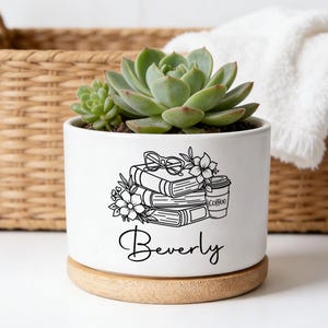 Op de afbeelding: Een witte cilindrische keramische plantenpot met een houten voet, met daarin een groene vetplant. De pot is voorzien van een zwarte lijntekening van gestapelde boeken, bloemen, een bril en een koffiebeker, met de naam Beverly in zwarte letters.