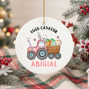 以下が含まれることがあります： 金色の紐が付いた、丸い白いセラミック製のクリスマスオーナメント。ウサギが運転するピンク色のトラクターが、ニンジンとイースターエッグを運んでいます。「EGGS-CAVATOR」と「ABIGIAL」の文字が入っています。