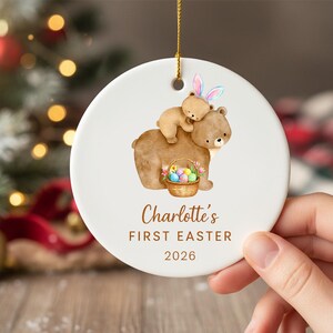 Puede incluir: Adorno navideño de cerámica blanca con una ilustración de acuarela de dos osos marrones. Uno de los osos lleva al otro, que lleva orejas de conejo. Se incluye una cesta de huevos de Pascua. El texto dice "Charlotte's FIRST EASTER 2026."