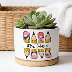 May include: A white ceramic planter with a succulent plant, featuring a pencil design and the name "Mrs. Johnson." The planter sits on a wooden base. A woven basket and a white towel are in the background.