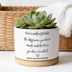 Può includere: Vaso in ceramica bianca con una pianta succulenta, con il testo "Never underestimate the difference you have made and the lives you have touched. Enjoy the next chapter." Il vaso poggia su una base di legno.