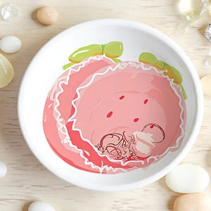 Puede incluir: Un cuenco de cerámica blanco con un diseño de fresa rosa. La fresa tiene lazos verdes y puntos rojos. El cuenco está rodeado de piedras decorativas y un anillo de diamantes.