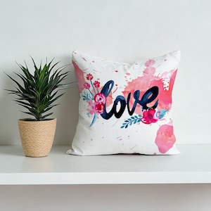 Puede incluir: Cojín cuadrado blanco con la palabra "love" en escritura azul marino, acentuado con salpicaduras de acuarela rosa y detalles florales. Un cojín decorativo para una cama o sofá.