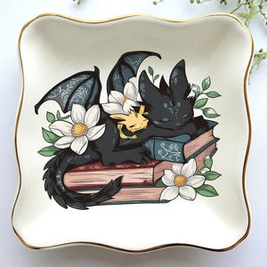 May include: Square ceramic trinket dish with a gold rim. The dish features a black dragon and a yellow creature sleeping on a stack of books, surrounded by white flowers and green leaves.