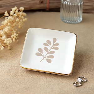 Op de afbeelding: Een vierkant, wit keramisch sieradenschaaltje met een gouden rand en een beige bladerontwerp. Het schaaltje staat op een bruin oppervlak, met twee zilveren ringen zichtbaar. Decoratieve droogbloemen en een glazen fles staan op de achtergrond.