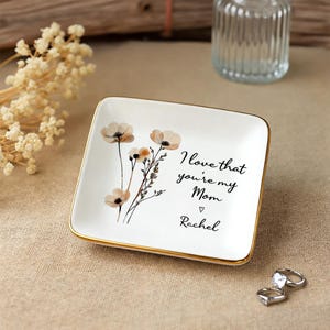 May include: Square white ceramic trinket dish with a gold rim, featuring a floral design and the text "I love that you're my Mom" with a heart and the name "Rachel".