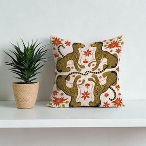 May include: A decorative throw pillow featuring a repeating pattern of stylized cheetahs and floral elements in shades of gold, black, and red on a cream background. The pillow is square and sits on a white shelf.