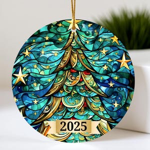 Op de afbeelding: Ronde kerstversiering met een glas-in-lood ontwerp met een gestileerde kerstboom in tinten van teal, goud en groen, versierd met gouden sterren. Het jaar "2025" staat op een banner. Een gouden touwtje is bevestigd om op te hangen.