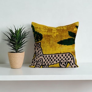 May include: A square cushion with a yellow background and a stylized animal design. The animal is beige with black spots. Dark green shapes are in the background. The cushion is on a white shelf.