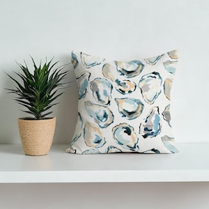 Puede incluir: Cojín cuadrado con un patrón de ilustraciones de conchas de ostra azules, grises y beige sobre un fondo blanco. A la izquierda del cojín hay una planta en maceta con hojas verdes.