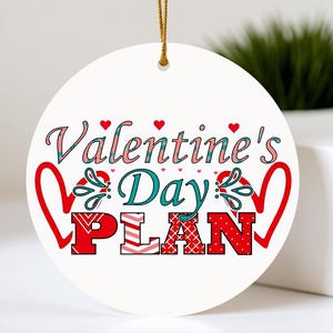May include: A white, circular Valentine's Day decoration featuring the text "Valentine's Day PLAN". The text is in red, teal, and pink, with heart motifs. It has a gold hanging string.