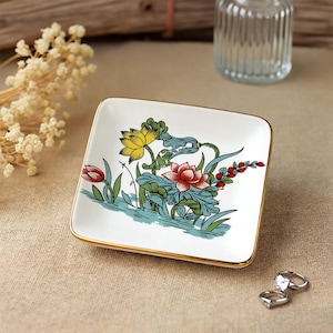 Può includere: Piatto quadrato in ceramica bianca con bordo dorato. Il piatto presenta un colorato motivo floreale con fiori gialli, rossi e blu. Probabilmente viene utilizzato per contenere piccoli gioielli.