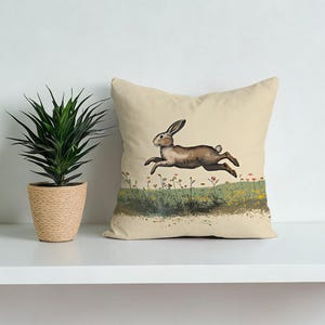 Könnte beinhalten: Ein beiges quadratisches Kissen mit einer Illustration eines springenden braunen Hasen. Das Kunstwerk zeigt den Hasen über einem Feld mit Wildblumen in Grün, Gelb und Rot. Das Kissen liegt auf einem weißen Regal neben einer Topfpflanze.
