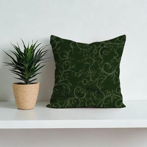 May include: A square, dark green decorative pillow with a repeating pattern of swirling vines and leaves in lighter shades of green. The pillow is placed on a white shelf next to a potted plant with green leaves and a woven beige pot.