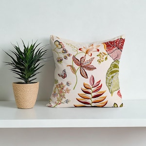 May include: A square decorative pillow with a cream-colored background. The pillow features a colorful floral pattern with red, orange, green, and pink leaves and flowers. A small butterfly is also visible. The pillow is placed on a white shelf next to a potted plant.