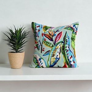 May include: Square throw pillow with a colorful botanical print. The design features large, stylized leaves in shades of blue, green, red, and orange against a light blue background. A potted plant sits to the left of the pillow.