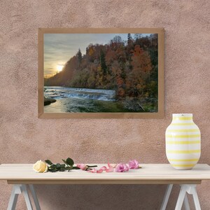 Könnte beinhalten: Gerahmte Landschaftsfotografie eines Wasserfalls und Flusses bei Sonnenuntergang. Das Bild zeigt eine Kaskade aus Wasser, Bäume mit Herbstlaub und einen goldenen Himmel. Eine gelb-weiß gestreifte Vase und Blumen stehen auf einem Tisch vor dem Foto.